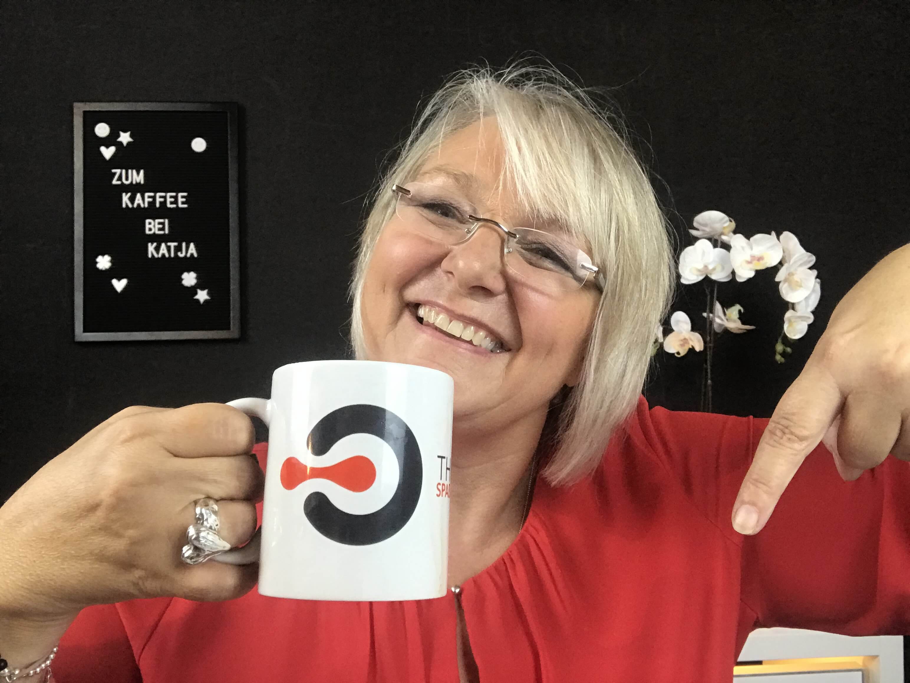 Katja Benny, l&auml;chelnd, mit Brille h&auml;lt eine Markentasse und zeigt darauf. Im Hintergrund sind eine kleine Tafel mit dem deutschen Text &bdquo;ZUM KAFFEE BEI KATJA&ldquo; und ein Orchideenarrangement zu sehen.