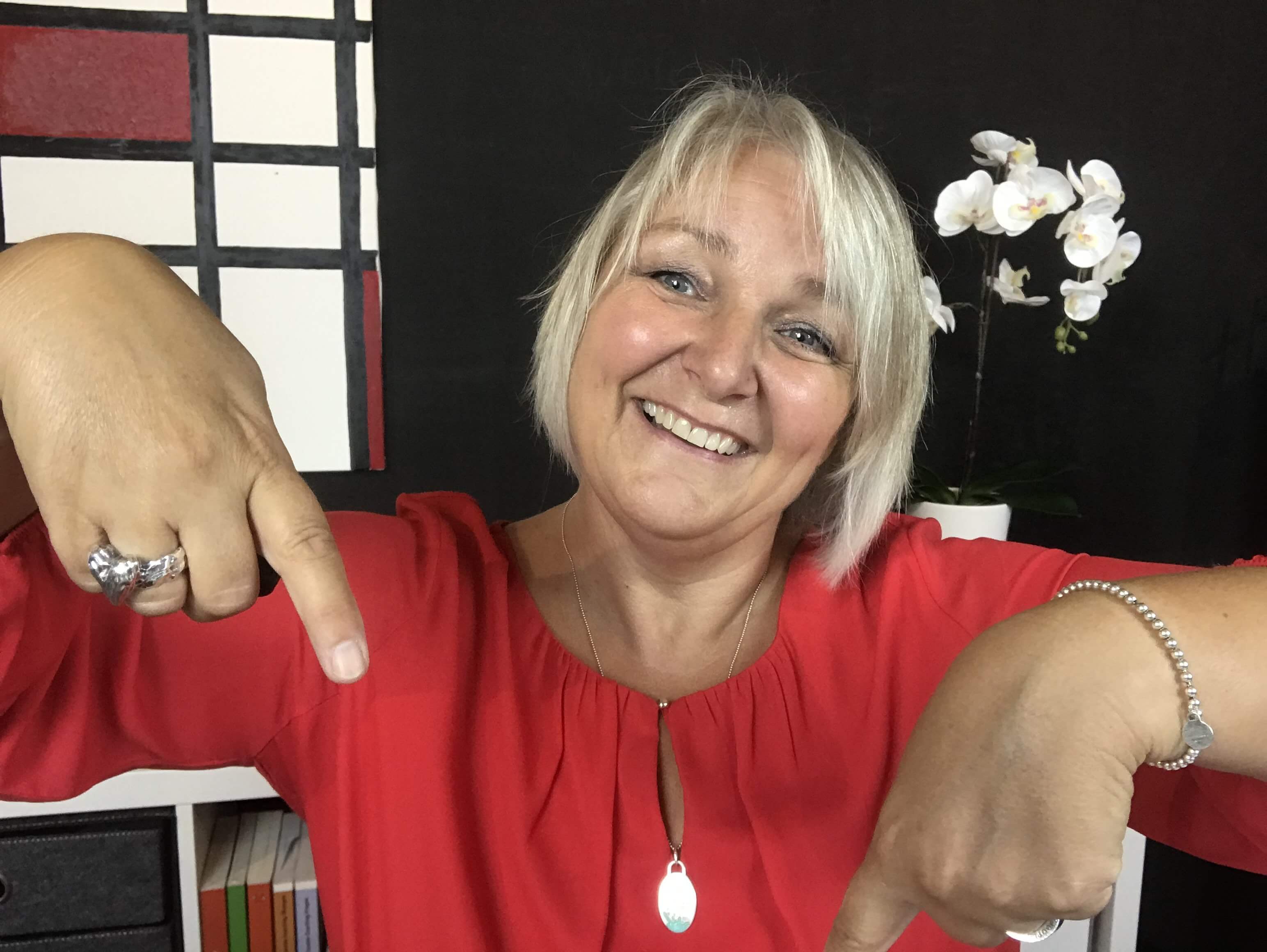 Eine Frau mit kurzen blonden Haaren in einer roten Bluse zeigt lächelnd mit beiden Händen nach unten. Im Hintergrund sind eine weiße Orchidee und Bücher zu sehen. Eine Frau mit kurzen blonden Haaren in einer roten Bluse zeigt lächelnd mit beiden Händen nach unten. Im Hintergrund sind eine weiße Orchidee und Bücher zu sehen.